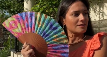 Une femme portant un haut orange tient un éventail multicolore avec des motifs géométriques, dans un cadre de jardin ensoleillé. Parfait pour l’été et les accessoires de mode