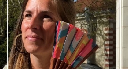 Une femme tient un éventail coloré avec des motifs géométriques, souriant sous le soleil. Idéal pour les accessoires de mode d’été.