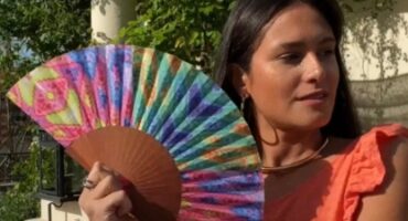 Une femme portant un haut orange tient un éventail multicolore avec des motifs géométriques, dans un cadre de jardin ensoleillé. Parfait pour l’été et les accessoires de mode