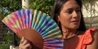 Une femme portant un haut orange tient un éventail multicolore avec des motifs géométriques, dans un cadre de jardin ensoleillé. Parfait pour l’été et les accessoires de mode