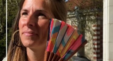 Une femme tient un éventail coloré avec des motifs géométriques, souriant sous le soleil. Idéal pour les accessoires de mode d’été.