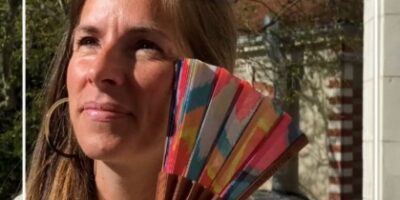 Une femme tient un éventail coloré avec des motifs géométriques, souriant sous le soleil. Idéal pour les accessoires de mode d’été.
